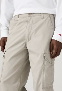 Bežové cargo nohavice vyrobené z jemnej látky, so šikmými a bočnými vreckami, zapínaním na gombík a prešitými detailmi na nohách.