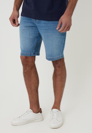Man draagt lichtblauwe denim shorts, een marineblauw overhemd en witte sneakers, staand tegen een effen achtergrond.