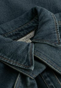 Gulerul jachetei din denim cu cusături vizibile și etichetă albă pe care scrie „NOELLA” și mărimea „S” în interiorul gâtului.