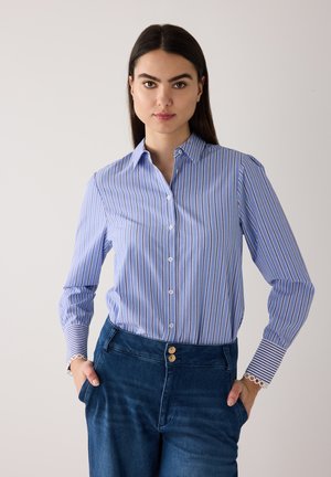 Frau mit dunklen Haaren, die ein blau-weiß gestreiftes Hemd mit Knöpfen und hoch taillierte, dunkelblaue Jeans trägt, die Hände in den Taschen.