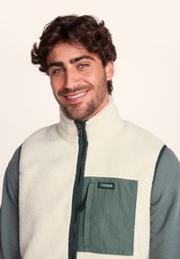 Fleece vest in cream with dark green accents. Features a high collar, front zipper, and a patch pocket on the left chest. Textured fabric.