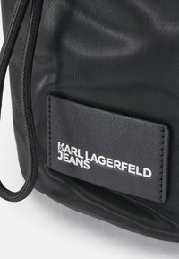 Borsa nera in nylon con finitura testurizzata, chiusura a coulisse e una patch rettangolare in pelle con la scritta "KARL LAGERFELD JEANS" in lettere bianche.