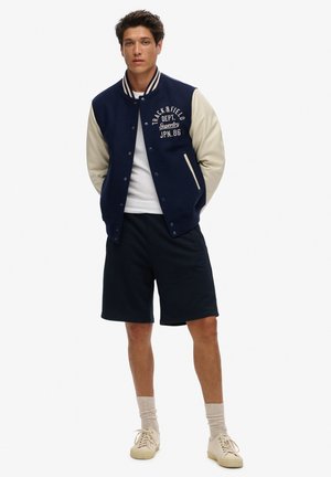 Chaqueta varsity de color marino con mangas crema, logo bordado en el pecho, camiseta blanca, pantalones cortos marinos y zapatillas beige. Texturas de tela suave.