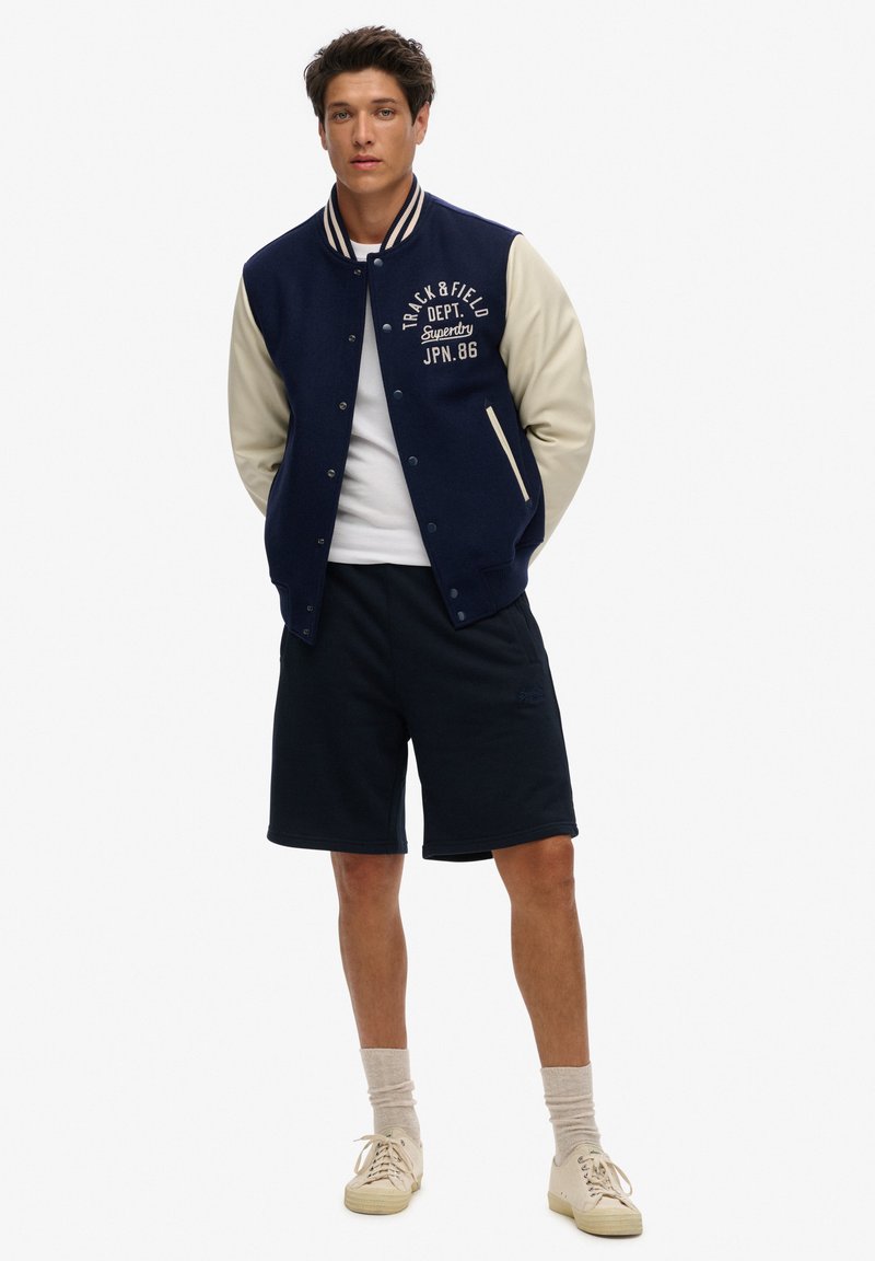 Giacca varsity blu navy con maniche crema, logo ricamato sul petto, t-shirt bianca, pantaloni corti blu navy e sneakers beige. Trame di stoffa morbide.