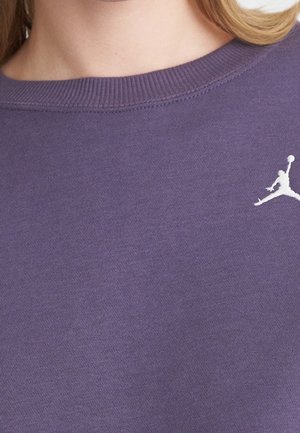 Zbliżenie osoby noszącej fioletową koszulkę z okrągłym dekoltem z małym białym logo koszykarza przy obojczyku.