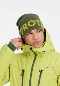 Grüne Mütze mit "PROTEST" in Gelb, kombiniert mit einer hellgrünen Jacke mit Reißverschlusstaschen und einer Kapuze mit Kordelzug.