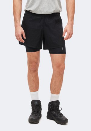 Homme portant un short de sport noir superposé, des chaussettes blanches, des chaussures de randonnée noires et un t-shirt gris, debout devant un fond uni.