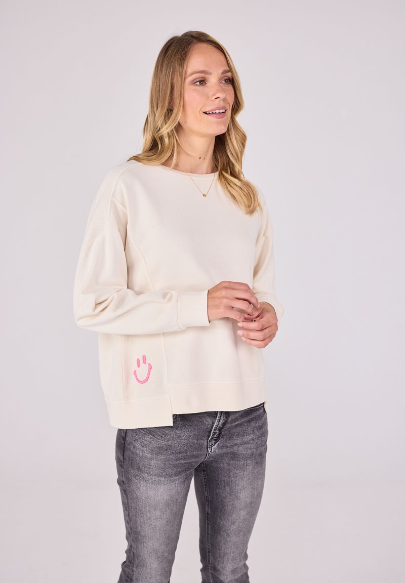 Helles Cream Sweatshirt mit lässiger Passform, ausgestattet mit einem rosa Smiley-Graphic auf der unteren linken Seite. Weicher Stoff und gerippte Bündchen.