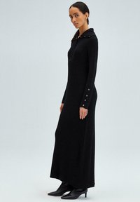 Maxi dress nera a coste con cappuccio, presenta una chiusura con zip e dettagli con bottoni sulle maniche. Abbinato a stivali neri con punta.
