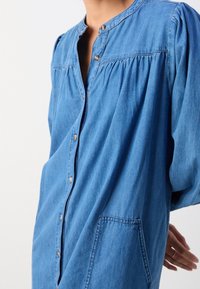 Denimskjorte med knappelukning foran, rund krave og puflangeærmer. Lys blå farve med en brystlomme og samlede detaljer.