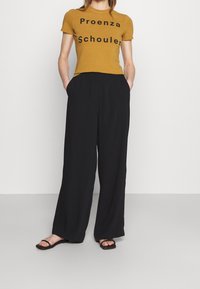 Senapsgul t-shirt med svart text "Proenza Schouler," kombinerad med svarta vida byxor och svarta platta sandaler.