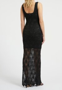 Nainen, jolla on päällä musta ilmanhihaa oleva virkattu mekko, jossa on läpinäkyvä hameosa ja paljastin, seisoo kirjoitettu pois valkoista taustaa vasten.