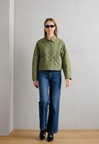Jachetă vesică, de culoare verde măslinie, cu două buzunare frontale, purtată peste blugi bleumarin evazați și cizme negre până la glezne, pe un fundal neutru.