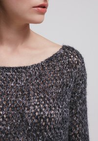 Grå stickad tröja med ett löst, öppet vävmönster, bred halsringning och texturerad yta. Har långa ärmar och en avslappnad passform.