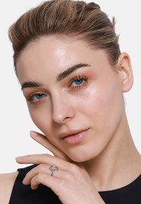 Zilveren ring met een verweven ontwerp, gedragen aan een vinger. Het model heeft een lichte huid en blauwe ogen, minimale make-up, neutrale achtergrond en een zwart topje.