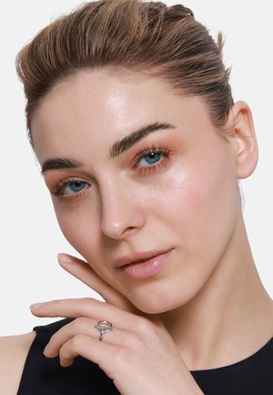 Silberner Ring mit verwobenem Design, am Finger getragen, Model hat helle Haut und blaue Augen, minimales Makeup, neutraler Hintergrund, schwarzes Oberteil.