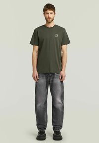 Donkergroene katoenen T-shirt met korte mouwen en een klein logo-opdruk, gecombineerd met vervaagde grijze jeans en zwarte schoenen. Effen achtergrond.