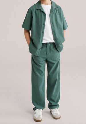 Person iført en grøn kortærmet skjorte med knapper, matchende grønne bukser, hvid T-shirt og hvide sneakers, stående med hænderne i lommerne.