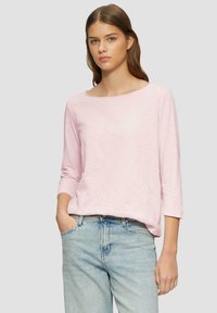 Haut rose clair à col bateau avec des manches trois quarts, fabriqué dans un tissu doux et texturé. Porté avec un jean en denim bleu clair taille haute.