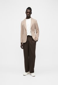 Άνδρας που στέκεται, φορώντας μπεζ σακάκι, λευκό πουκάμισο, σκούρο καφέ παντελόνια με φαρδιά μπατζάκια και λευκά αθλητικά παπούτσια, μπροστά σε απλό λευκό φόντο.