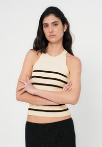 Kremfarvet ribbet tanktop med vandrette mørkebrune striber, med en tætsiddende pasform og halterhalsudskæring. Sort blonder nederdel nedenunder.