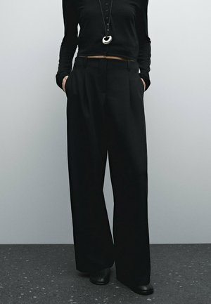 Personne portant un pantalon noir ample, un haut noir à manches longues et des chaussures noires, les mains dans les poches, avec un collier pendentif circulaire.