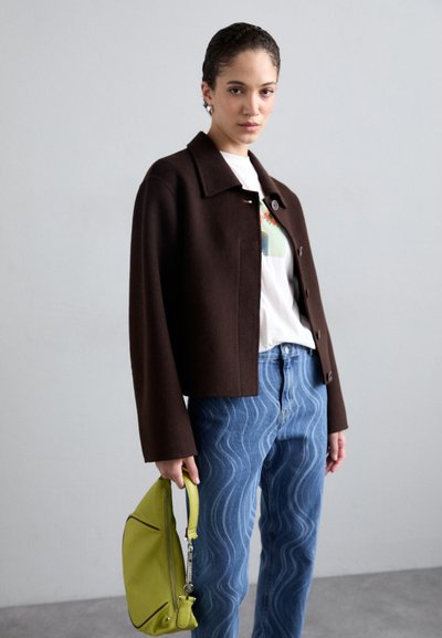 Brun cropped jakke med knappelukking, hvit t-skjorte med grafikk, blå jeans med bølgemønster, og en grønn håndveske.