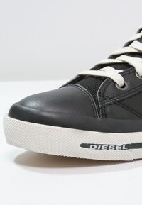 Schwarze Leder-Sneaker mit weißer Gummisohle, texturiertem Zehenkappen und kontrastierenden cremefarbenen Schnürsenkeln; mit "DIESEL"-Markierung entlang der Seite.