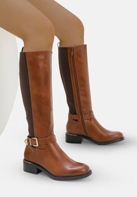 Botas marrones hasta la rodilla hechas de cuero sintético liso, con cremallera lateral, panel de tela texturizada en marrón y una hebilla decorativa. Tacón bajo.