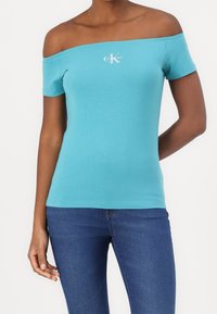 Top azul claro con hombros descubiertos, hecho de una tela suave, con mangas cortas y un logo blanco de Calvin Klein en el frente.