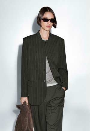 Blazer scuro a righe con una vestibilità rilassata, abbinato a un top grigio e pantaloni coordinati. La modella tiene in mano una giacca in suede marrone. Sono presenti occhiali da sole.