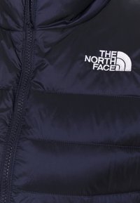 Marineblauer Puffer-Jacke mit gestepptem Muster, durchgehendem Reißverschluss und Kapuze. Verfügt über ein weißes, gesticktes Logo auf der oberen linken Seite.