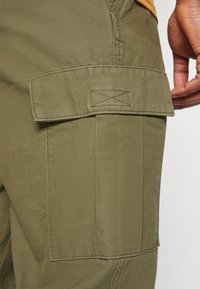 Olijf groene cargobroeken hebben een grote klepzak aan de zijkant, gemaakt van stevig katoenmateriaal met zichtbare stikdetails.