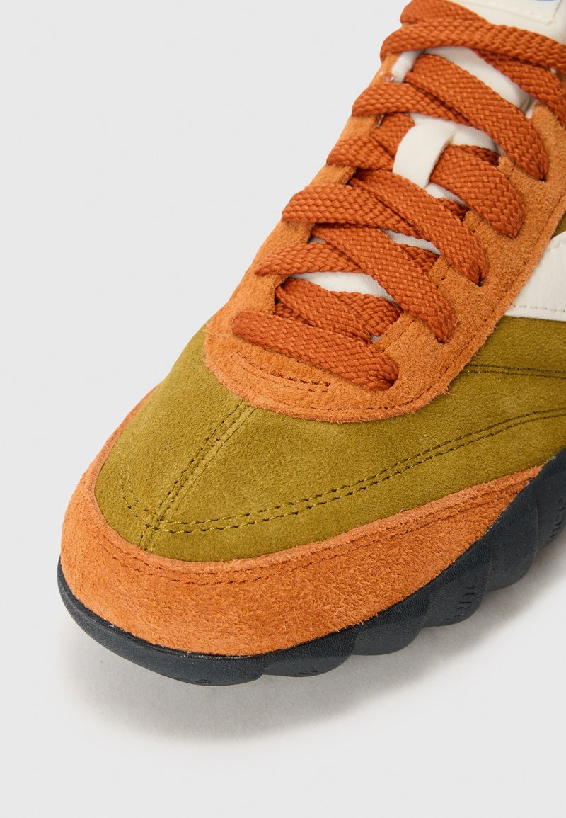 Zapatilla de ante naranja y oliva con puntera naranja y cordones naranjas, suela negra texturizada, sobre un fondo blanco.
