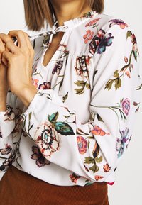 Blusa floreale in tessuto bianco, con motivi di fiori colorati, scollo volante, dettaglio a chiave e maniche elasticizzate.