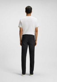 Camicia bianca da uomo a maniche corte con finiture a contrasto, abbinata a jeans neri a gamba dritta e scarpe scure, vista da dietro.
