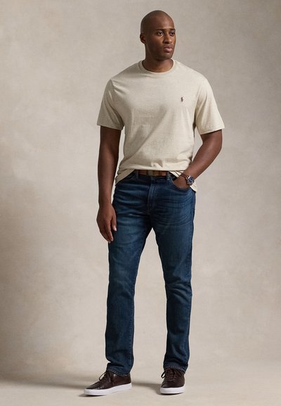 Beige bomullst-shirt med logo på bröstet, kombinerad med mörkblå denimjeans och bruna läderskor. Enkel design, ledig passform.