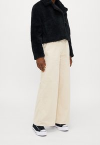 Schwarze, cropped Kunstfelljacke, cremefarbene, weit geschnittene Cord-Hosen und schwarze Sneakers mit niedrigen Schafthöhen und weißen Sohlen. Glatte Texturen und gegensätzliche Farben.