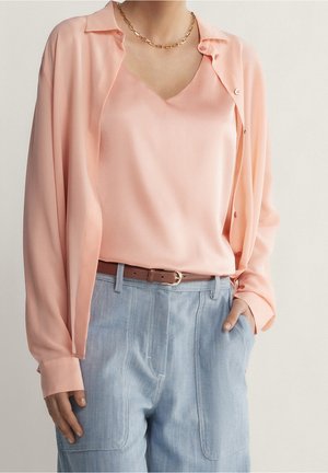 Frau trägt eine hellrosa Bluse und ein Top, in hellblaue, hoch taillierte Hosen mit braunem Gürtel gesteckt, sichtbare goldene Kette.