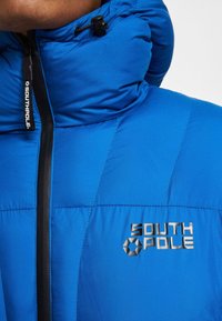 Kék puffos dzseki magas gallérral, látható cipzárral és fekete díszítésekkel. A mellkason ezüst színű "SOUTH POLE" logó látható.