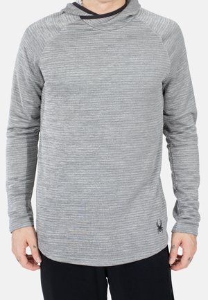Homme portant un hoodie gris à manches longues rayé avec un petit logo araignée noir en bas à gauche, associé à un pantalon noir.
