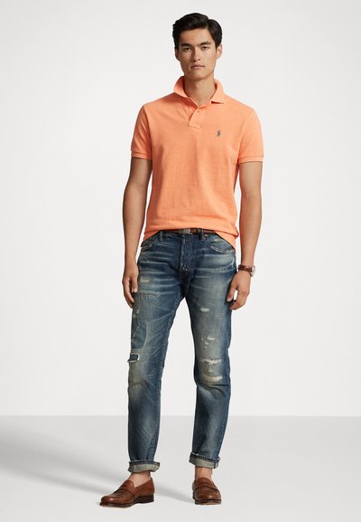 Bărbat îmbrăcat cu un tricou polo portocalie, blugi albaștri distruși cu manșete răsucite, mocasini din piele maro și un ceas de mână, stând pe un fundal simplu.