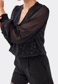 Blouse noire avec des manches transparentes, des accents métalliques à pois, un décolleté en V et des poignets froncés, assortie à un short noir ajusté.