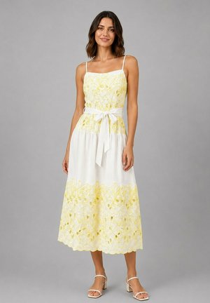 Femme portant une robe blanche sans manches avec une broderie florale jaune et une ceinture à la taille, debout devant un fond gris uni.