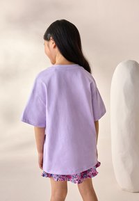 Fliederfarbenes Baumwoll-T-Shirt mit kurzen Ärmeln, lockerer Passform und rundem Ausschnitt, kombiniert mit einem geblümten, gerafften Rock. Glatte Textur, lässiges Design.