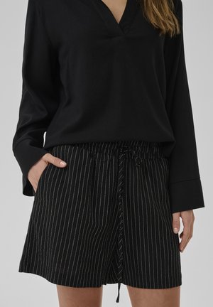 Kvinde iført sort langærmet V-hals top og sorte shorts med hvide lodrette nålestriber, den ene hånd i lommen mod en grå baggrund.