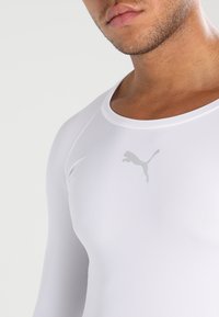 Chemise de sport à manches longues blanche en tissu lisse et extensible. Présente un logo Puma gris discret sur la poitrine et un col rond.