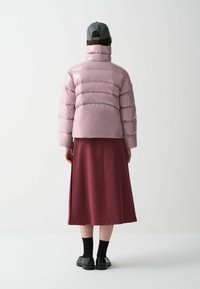 Ljusrosa puffjacka med horisontella kviltade sektioner, hög krage, parat med en vinröd kjol, svarta strumpor och svarta skor.