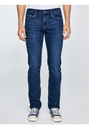 Muž nosící tmavě modré úzké džíny a námořnicky modré plátěné tenisky stojící před jednobarevným světlým pozadím.