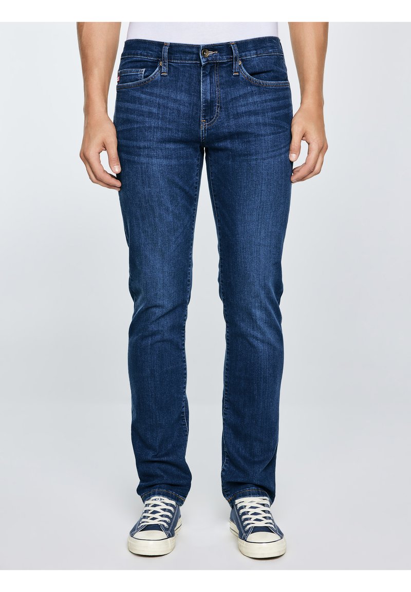 Muškarac u uskim tamnoplavim trapericama i mornarskoplavim platnenim tenisicama stoji naslonjen na jednobojnu svijetlu pozadinu.